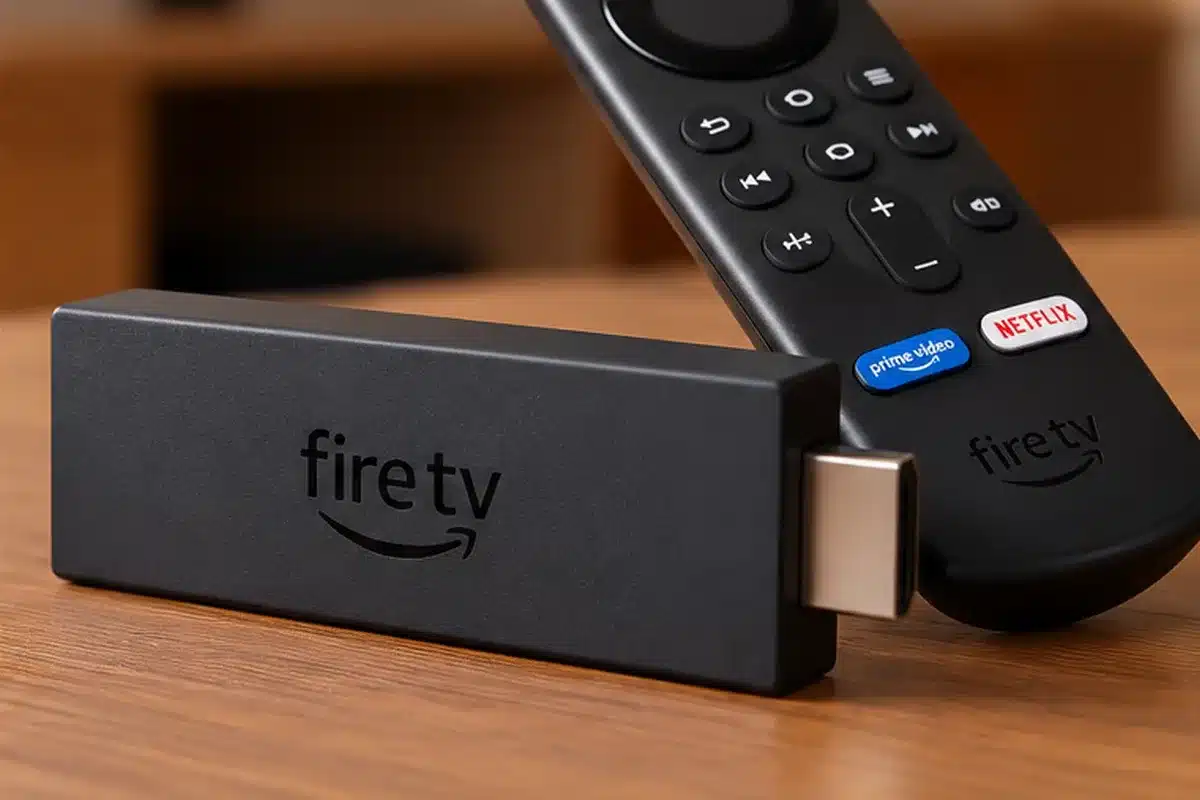 Dispositivo de streaming Fire TV Stick preto com controle remoto, sobre mesa em cenário de sala com televisão desfocada